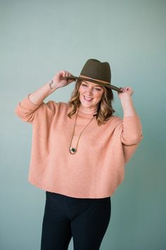 a woman wearing a brown hat and black pants with her hands on her head, smiling