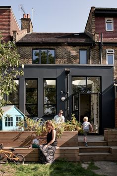 a woman and two children are sitting on the steps in front of a house with black walls