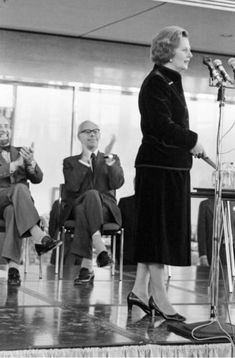 a woman standing at a podium with microphones in front of her and other people