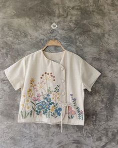 a white shirt with flowers on it hanging from a wooden hanger against a gray wall