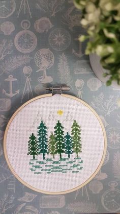 a cross stitch christmas scene with pine trees and snowflakes on the wall next to a potted plant