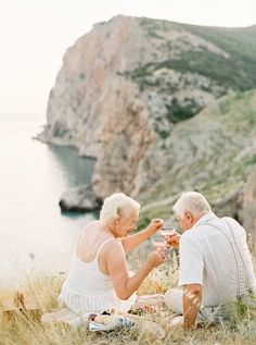 an older couple sitting on the grass with wine in their hands and looking at each other