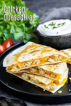 chicken quesadillas on a black plate with sour cream and tomatoes in the background