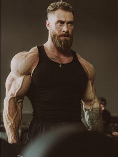 a man with a beard and tattoos standing in front of a mirror wearing a black tank top