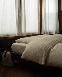 an unmade bed in front of two windows with white curtains