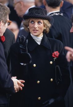 a woman in a black coat and hat talking to two men wearing red roses on her lap