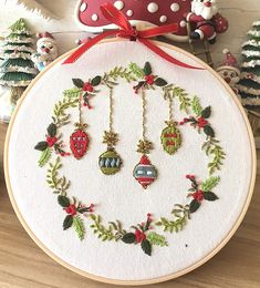 a cross stitch christmas wreath with ornaments on it and a red ribbon hanging from the hoop