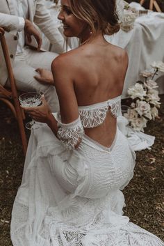 the back of a woman in a white wedding dress holding a glass of wine while sitting down