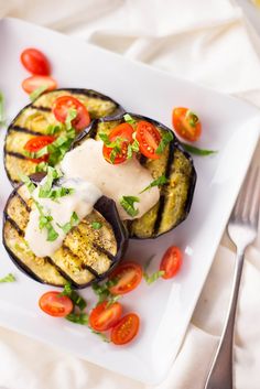 grilled eggplant with tomatoes and dressing on a white plate next to a fork