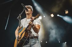 a man holding a guitar while standing in front of a microphone on stage with lights behind him