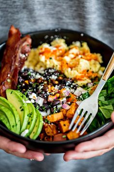 a person holding a black bowl filled with vegetables and bacon, avocado, and eggs