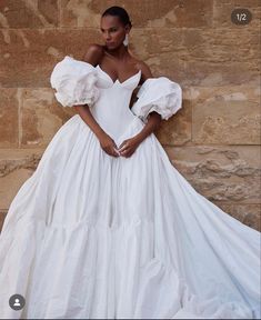 a woman in a white wedding dress leaning against a stone wall with her hands on her hips