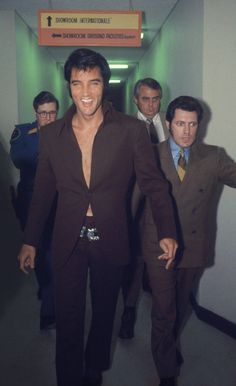 a man in a suit walking down a hallway with his arms out and two other men behind him