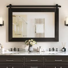 a bathroom vanity with two sinks and a large mirror above it, in front of a wall mounted towel dispenser