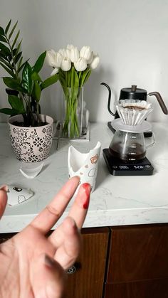 a person is pointing at some flowers on a counter top with a coffee maker in the background