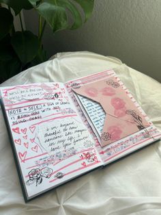 an open book with pink and white designs on it sitting on a bed next to a potted plant