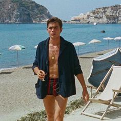 a shirtless man walking on the beach next to an umbrella and chair with water in the background