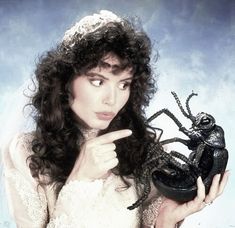 a woman in a wedding dress holding a black beetle on her finger and pointing to the camera