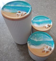 three cups with sand and shells painted on them sitting on the floor next to each other