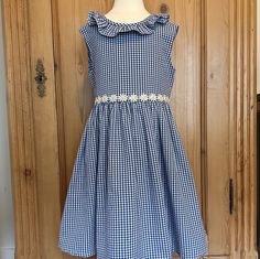 "Beautiful sleeveless gingham dress made from navy and white medium weight 100% cotton. Classic design with ruffle embellishment at collar that extends to v shaped back neckline, lined bodice, back zipper and self fabric ties with back bow. Adorned with white and yellow daisy trim around the waist. Perfect summer party or special occasion dress. Size 5 from the pattern. Please measure a well fitting dress for proper fit due to the variability of sizing in patterns of different vintage and style. width at chest is 14\"/35 cm waist to hem length 16\"/40cm Can purchase with a little growing room and cinch with waist ties. All my dresses are one of a kind. Bespoke dresses in this style can be obtained in sizes 2-5." Fitted Gingham Sleeveless Sundress, Cotton Lace Trim Dress For Picnic, Gingham Dress With Smocked Back For Garden Party, Sleeveless Gingham Dress With Ruffles, Gingham Cotton Dress For Garden Party, Navy Sleeveless Dress With Ruffles, Cotton Gingham Dress With Lace Trim, Gingham Cotton Dress With Lace Trim, Picnic Gingham Dress With Lace Trim