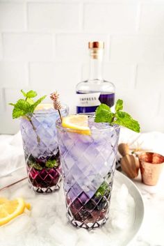 two glasses filled with purple and green drinks on top of a marble table next to a bottle