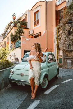 a woman standing next to an old car in front of a pink and orange building