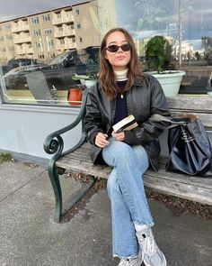 a woman sitting on a bench in front of a window holding a book and wearing sunglasses