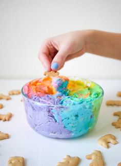 a child's hand dips colored food into a bowl with cookies on the side