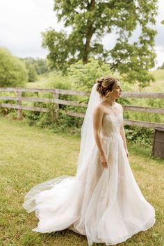 a woman in a wedding dress standing on the grass near a fence and trees, looking off into the distance