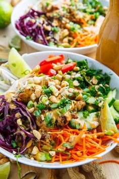 two white bowls filled with colorful salad and dressing on top of a wooden table next to spoons