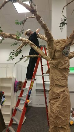 two people on ladders working on a fake tree in an office setting with cardboard covering the branches