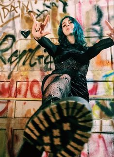 a woman with blue hair and black dress posing in front of a wall covered in graffiti