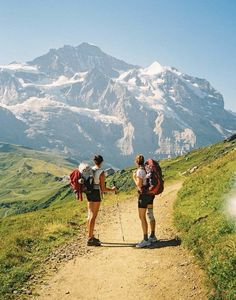 two people walking up a dirt path in front of a mountain range with snow capped mountains behind them