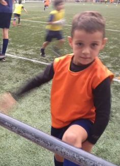 a young boy holding onto a metal pole on a soccer field with other children in the background