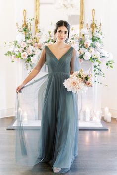 a woman standing in front of a fireplace wearing a long dress and holding a bouquet