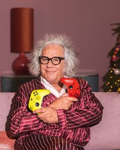 an old woman sitting on a couch holding two video game controllers in one hand and a controller in the other