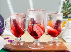 three glasses filled with liquid sitting on top of a wooden cutting board next to purple flowers