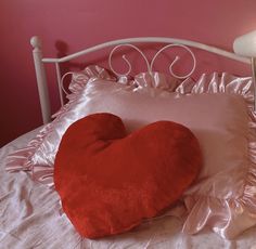 a heart shaped pillow on top of a white bed with pink sheets and a lamp next to it