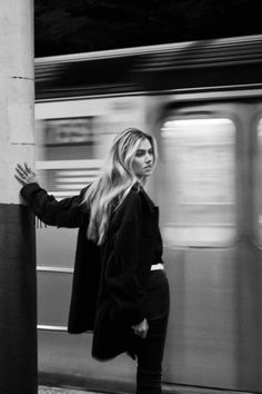 a woman standing next to a pole in front of a subway train as it passes by