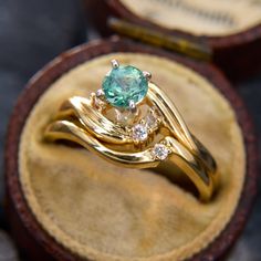 two gold rings with green and white stones on them sitting in a wooden ring box