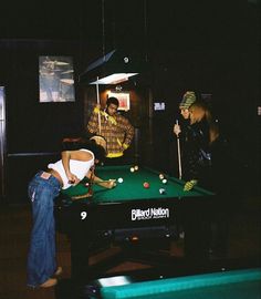 some people are playing pool in a dark room with lights on and one person is leaning over the pool table