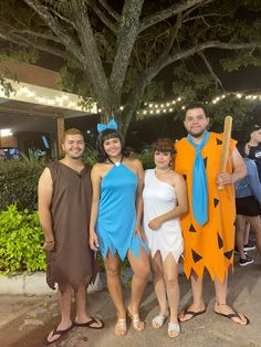 four people dressed up in costumes posing for a photo at an outdoor event with lights strung from the trees