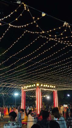 people are standing under string lights at night in an open area with red pillars and white columns