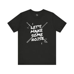 A graphic T-shirt featuring a design of drumsticks and the phrase 'Let's Make Some Noise'. This T-shirt is perfect for music lovers, drummers, and anyone who loves to stand out with a unique style. It gives off a cool, edgy vibe and is great for casual wear or music events. Perfect for birthdays, music concerts, and everyday wear. The available colors are listed in the dropdown menu. You can refer to the color chart to see those colors. Not all colors on the color chart are available for each de Band Merch Tri-blend Graphic T-shirt, Music-themed Graphic Short Sleeve Top, Music-themed Graphic Design Short Sleeve Top, Music-themed Slogan T-shirt With Short Sleeves, Music-themed Slogan Tops For Concerts, Music-themed Slogan T-shirt For Concert, Band Merch Slogan Shirt For Fans, Band Merch Shirt With Slogan For Fans, Band Merch Shirt With Slogan