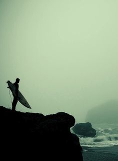 a man holding a surfboard standing on top of a cliff next to the ocean