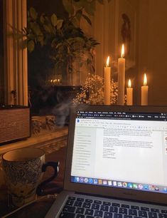 an open laptop computer sitting on top of a desk next to a candle lit window