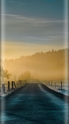 an empty road with snow on the ground and trees in the background at sunrise or dawn