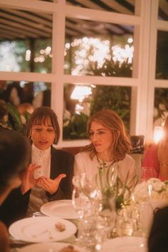 three people sitting at a table talking to each other in front of a window filled with lit candles