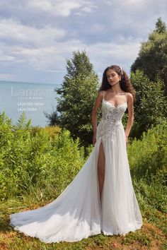 a woman in a white wedding dress standing on the grass by the water with trees