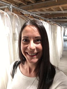 a woman is smiling in front of some white dresses hanging on a rack and looking at the camera